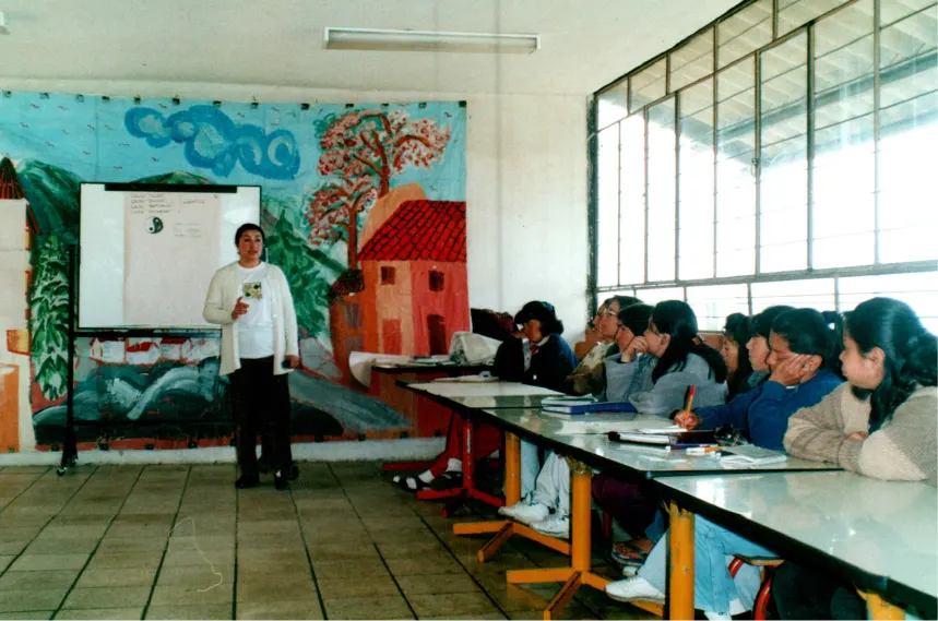 Docente del INEPE impartiendo una clase a un grupo de estudiantes en un aula con mural artístico, reflejando los inicios del proyecto educativo comunitario.