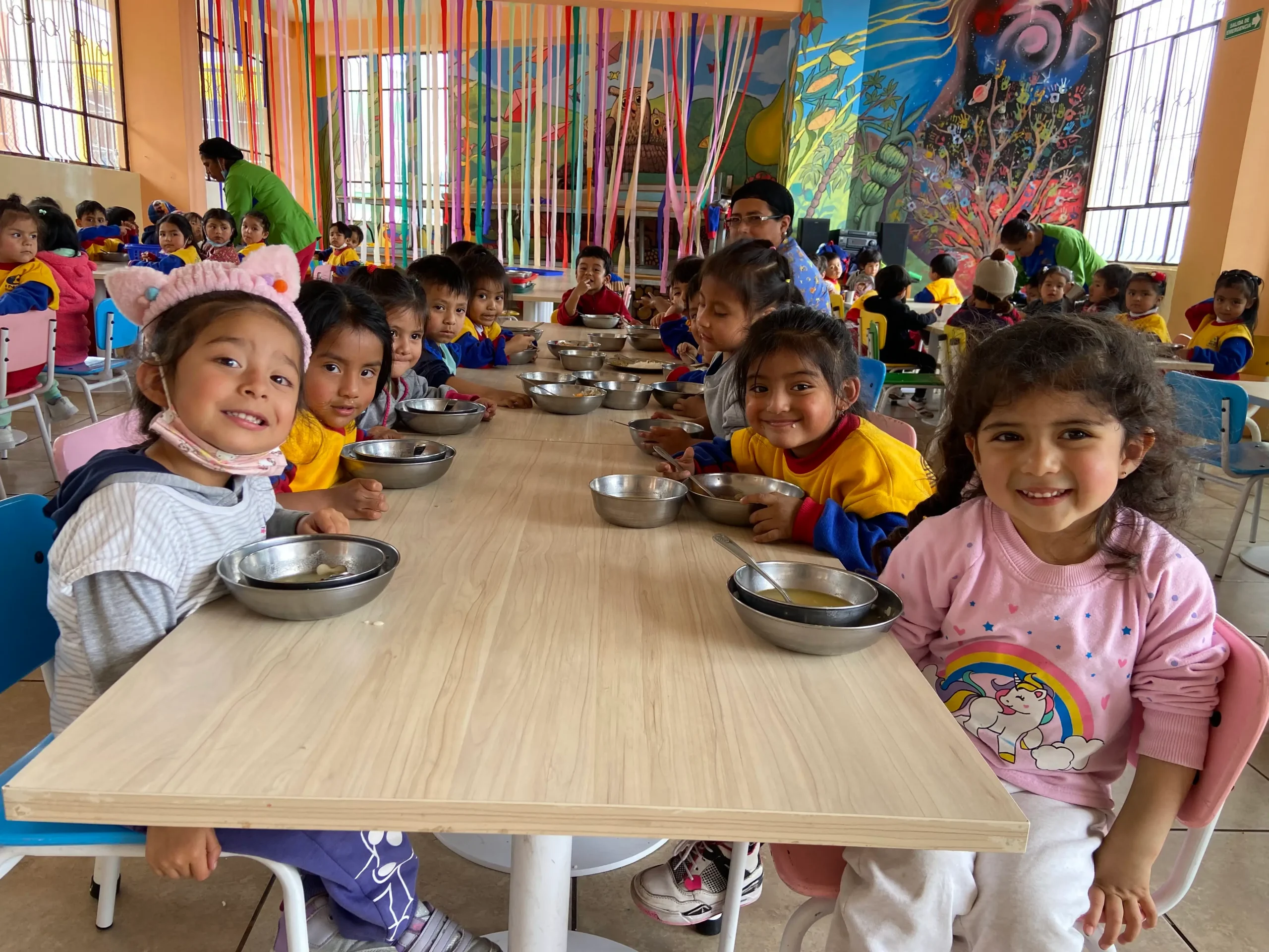 Niños y niñas del INEPE sonriendo mientras disfrutan de un almuerzo saludable en el comedor comunitario escolar, rodeados de murales artísticos.