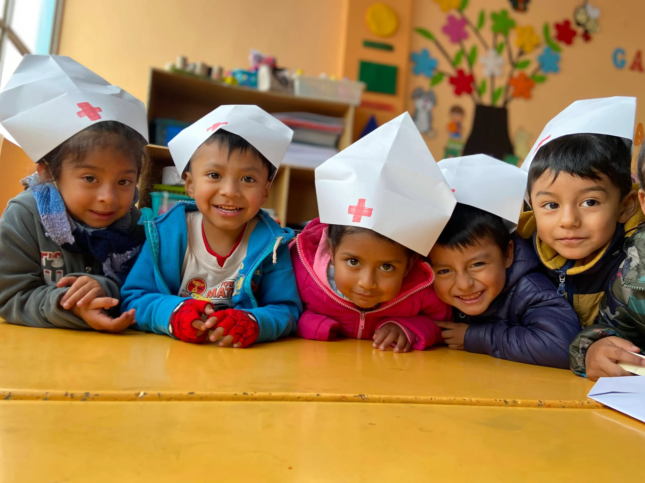 Niños y niñas del INEPE sonriendo con gorros de enfermería de papel, invitando a las personas a unirse al programa de apadrinamiento escolar.