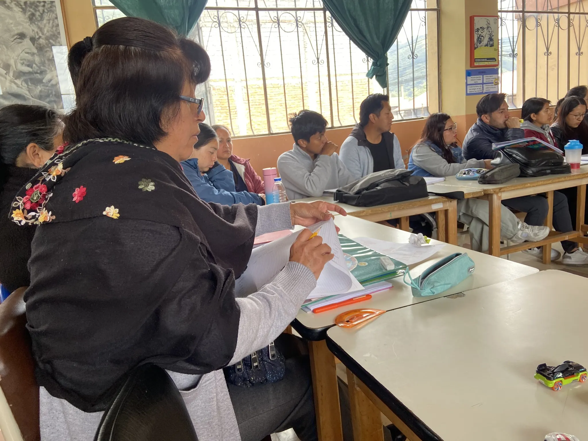 n primer plano, una mujer con lentes y un chal negro bordado revisa documentos sobre un pupitre escolar. Al fondo, varios hombres y mujeres adultos escuchan atentamente durante una reunión en un aula iluminada.