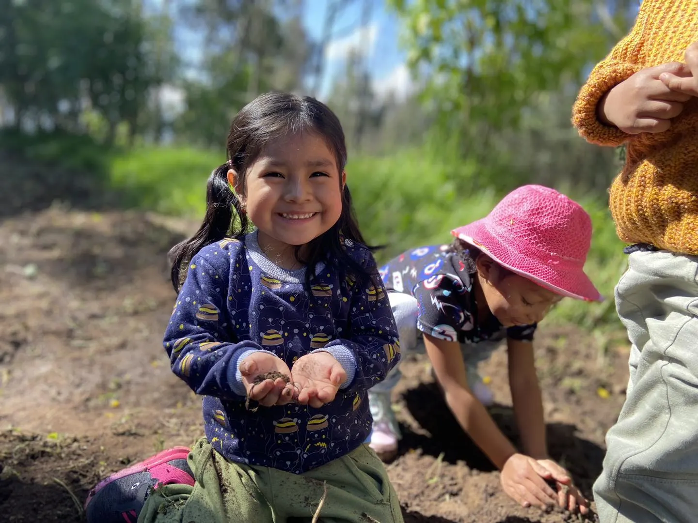 Niña pequeña sonriendo a la cámara con las manos llenas de tierra, arrodillada en un huerto escolar junto a otros compañeros durante una actividad de siembra.