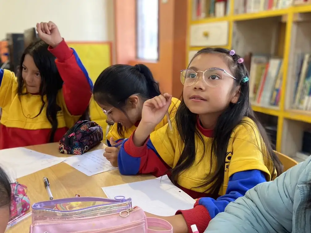 Primer plano de una estudiante prestando atención mientras su compañera levanta la mano para participar. El INEPE promueve un ambiente de aula donde la curiosidad, la pregunta y la escucha activa son esenciales para el aprendizaje significativo.