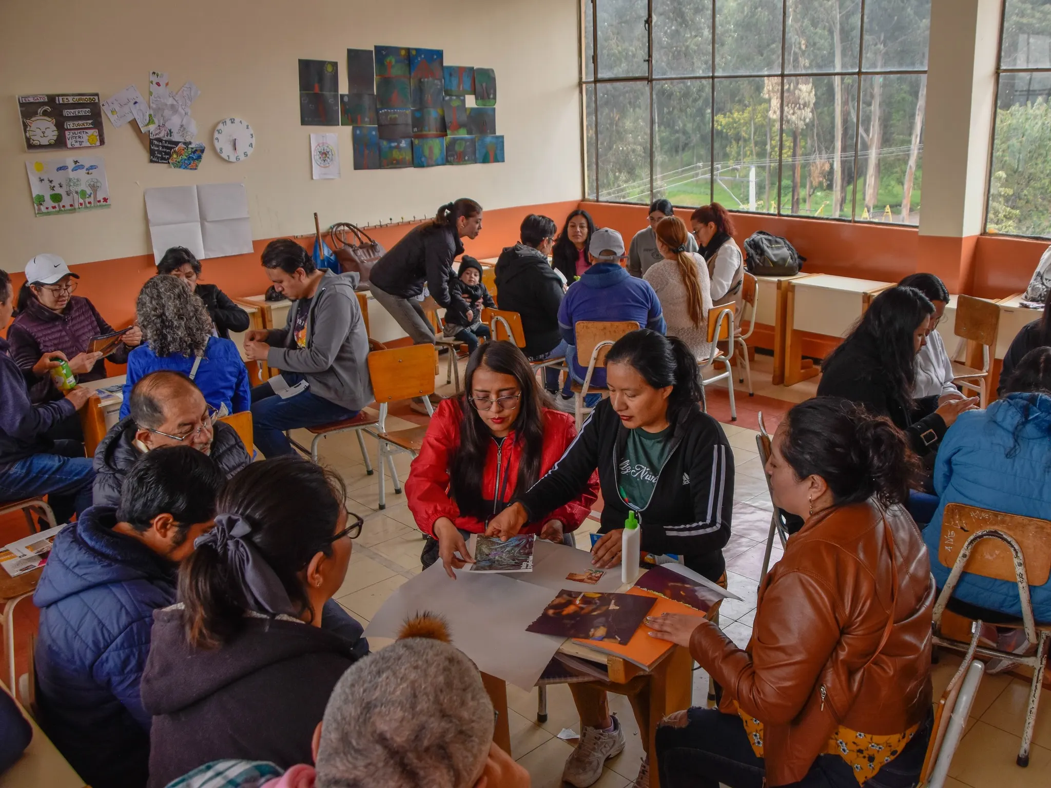 Varios grupos de adultos sentados alrededor de mesas escolares en un aula iluminada, trabajando con recortes, pegamento y cartulinas durante una actividad participativa, con dibujos infantiles pegados en las paredes de fondo.