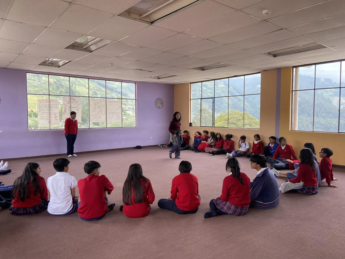 Un grupo numeroso de estudiantes adolescentesestán sentados en un gran círculo en el suelo de un salón amplio. Una Docente está de pie en el centro del círculo hablando, mientras grandes ventanales muestran un paisaje montañoso verde de fondo.