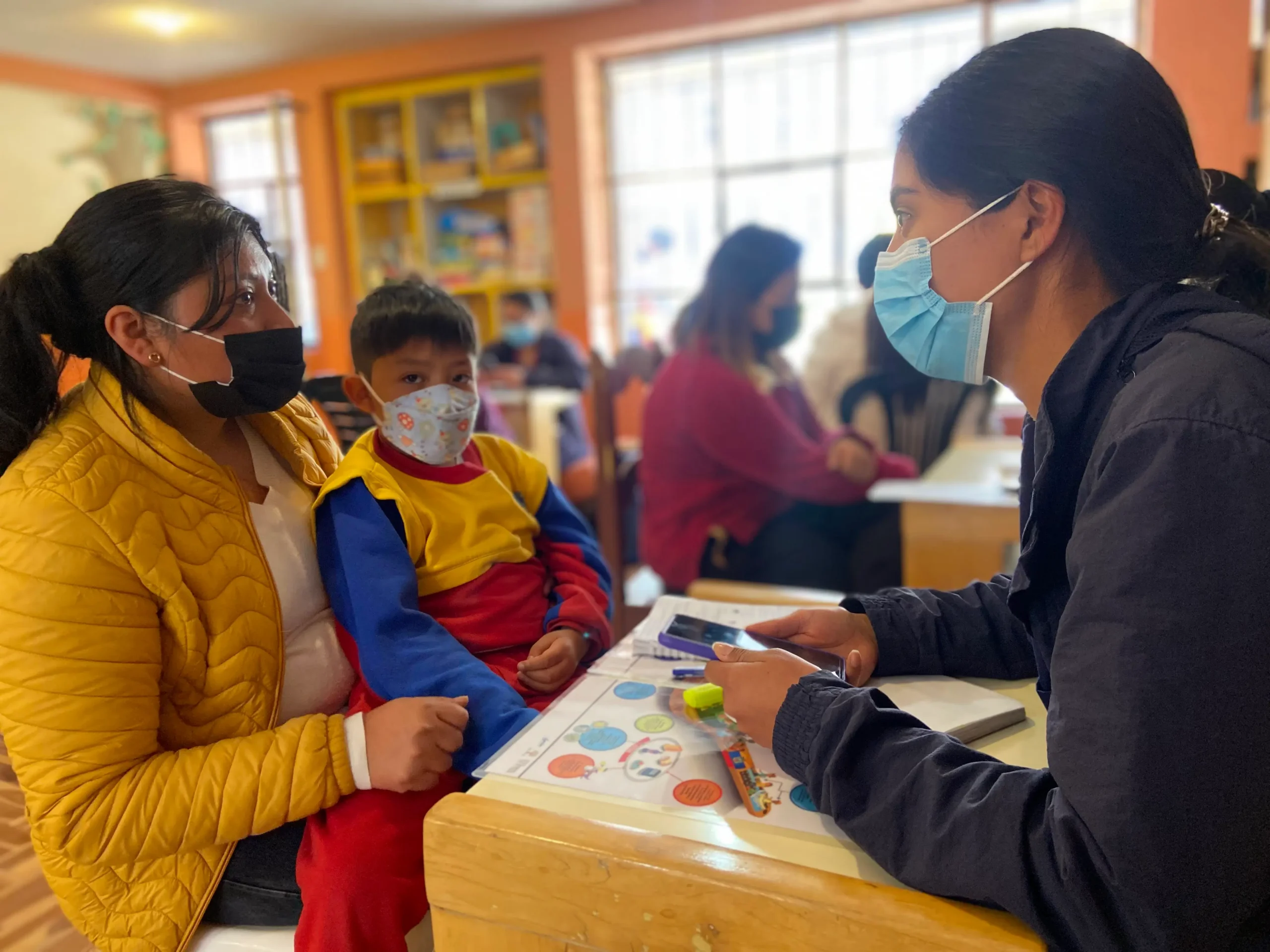 Una mujer con mascarilla azul conversa con una madre y un niño pequeño sentados a una mesa en una oficina de atención.